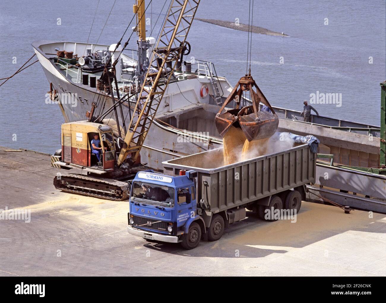 Historical 1980s archive aerial view of empty haulage contractor tipper truck reversed up to open hold of a docked bulk carrier freighter ship with dock worker driving 80s crane unloading dusty grain into lorry at dockside on Mistley Quay a small port & dock business on River Stour an archival image of the way we were in 1986 at Manningtree Essex England UK Stock Photo