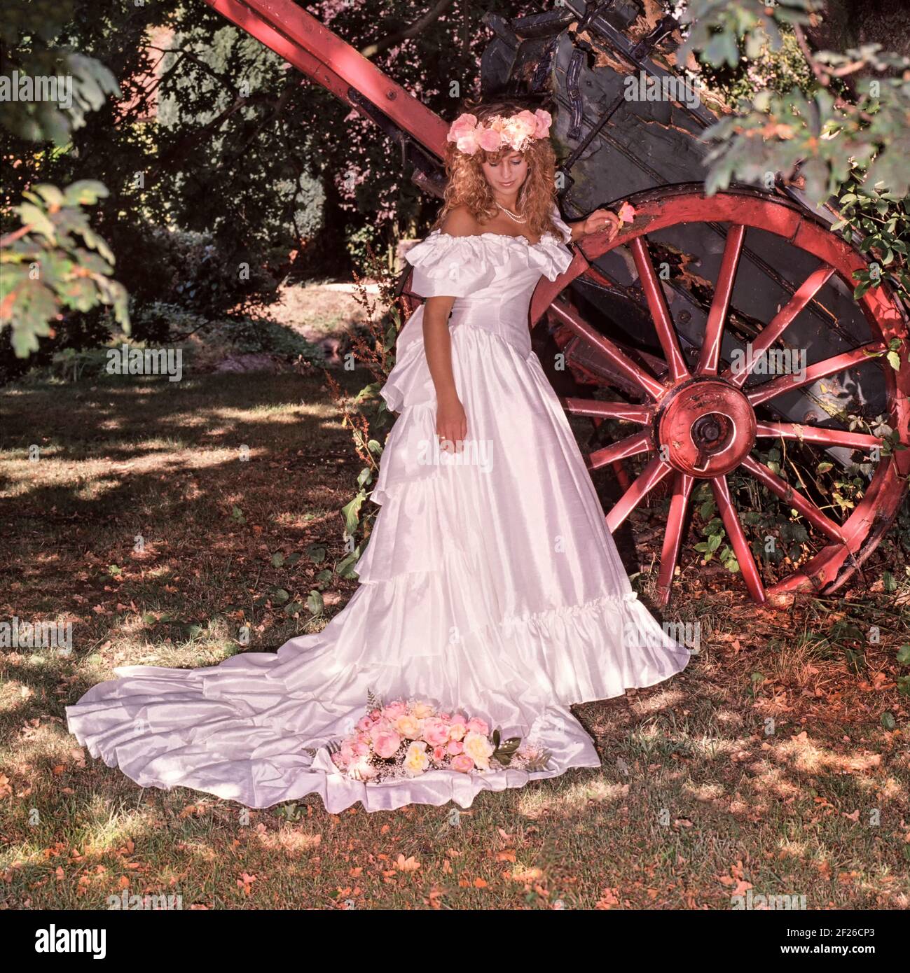 1980s historical archive view of 1989 young model released bride posing for photograph outdoors beside cartwheel prop wearing full length off the shoulder 80s wedding dress and train in colour photo with bouquet & matching headdress archival fashion image of the way we were in 1900s Essex England UK Stock Photo