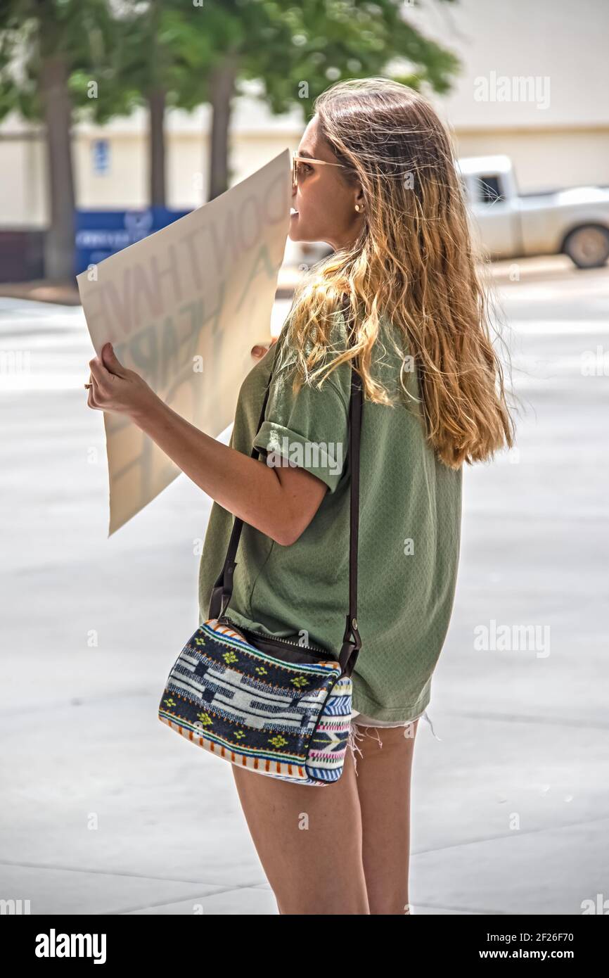 7 2 2019 Tulsa USA - Attractive tanned woman in short shorts and sunglasses with long brunette hair and cute purse stands by street with protest sign Stock Photo