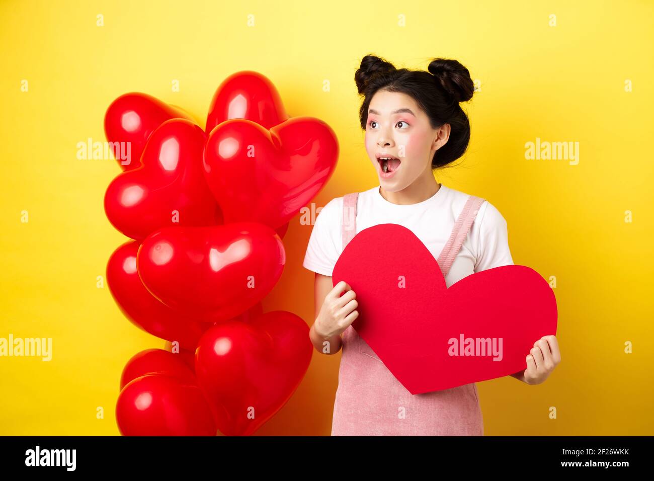 Happy Valentines day. Excited and surprised asian girl checking out romantic offer, looking left cheerful, showing big red heart Stock Photo