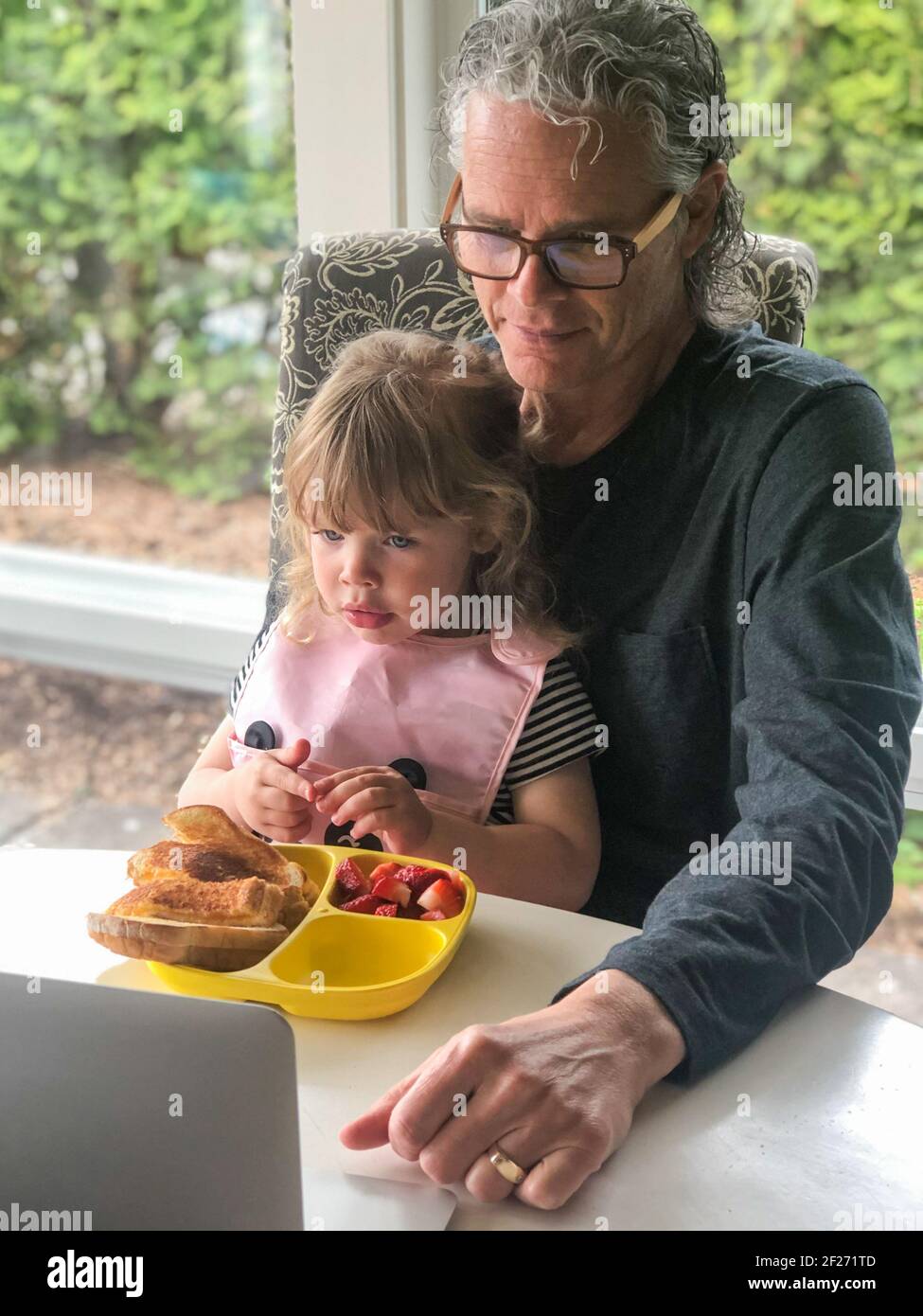 Caucasian grandfather and his small granddaughter learning using a laptop while eating lunch. Family multi-generation leisure activities. Family time. Stock Photo