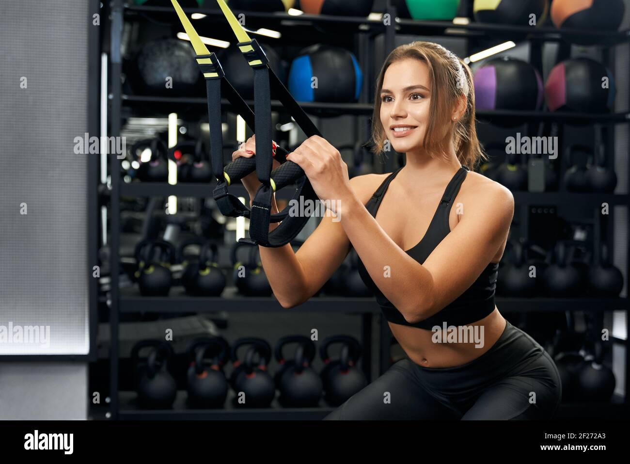 Close up of attractive young fitness woman doing cardio exercise with TRX system in modern gym. Concept of fitness straps and special sports equipments for workout in gym. Stock Photo