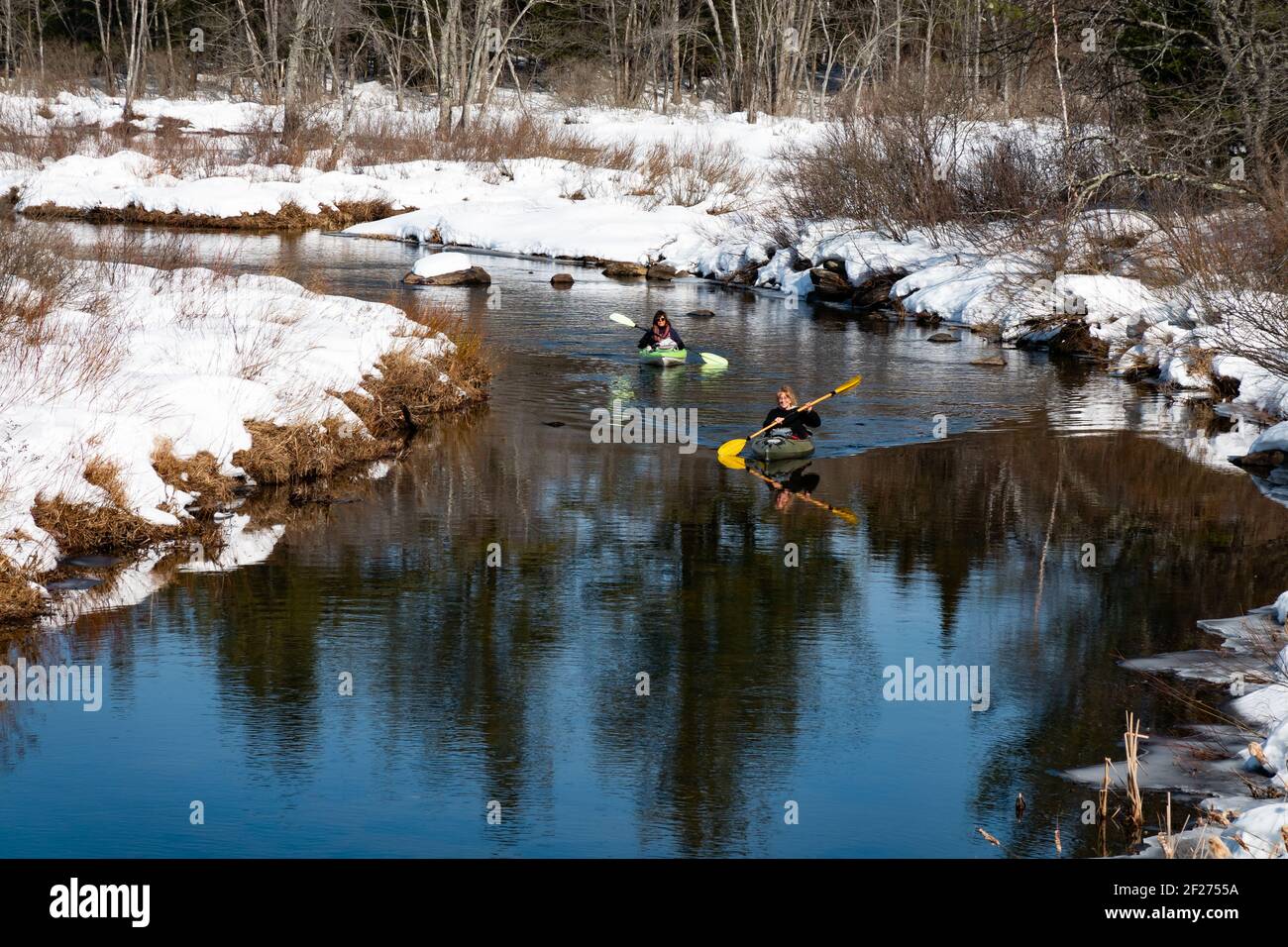 Two mature women paddling kayaks on the Kunjamuk River near Speculator, NY USA in the Adirondack Mountains in late winter with snow on the riverbanks Stock Photo