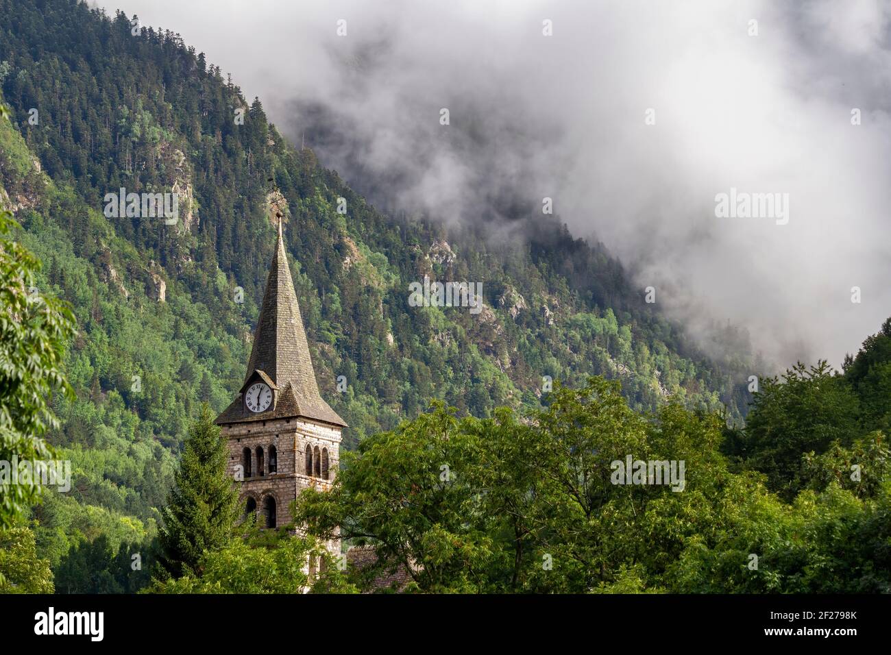 bell tower with a clock in a mountain village. Clouds cover a hillside in the background, in Arties, Valle de Aran Lleida, Spain Stock Photo