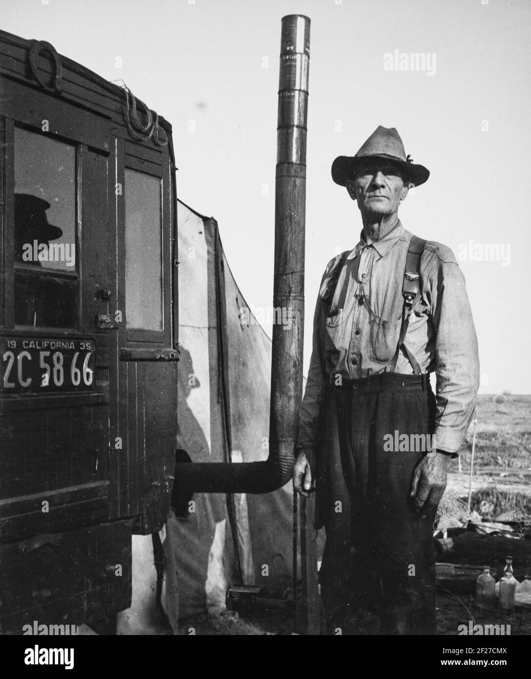 Photograph shows a man, three-quarter length portrait, standing next to tent dwelling with chimney, possibly attached to motor vehicle. . 1935. Photograph by Dorothea Lange Stock Photo