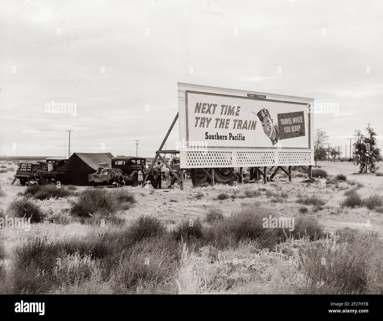 Next Time Take The Train billboard. Three families camped on the plains along U.S. 99 in California. They are camped behind a billboard which serves as a partial windbreak. All are in need of work. November 1938. Photograph by Dorothea Lange Stock Photo