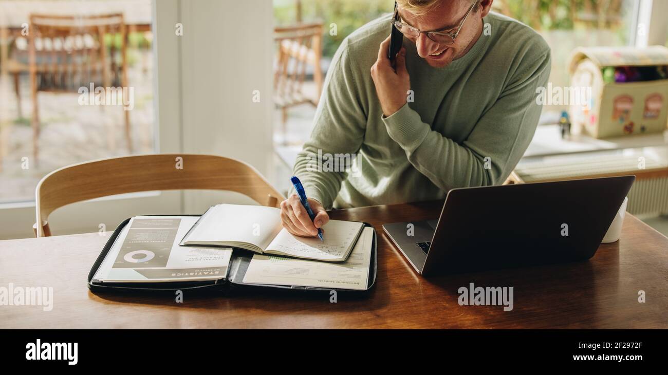 Man sitting at table talking on phone and writing in a book. Man working from  home with laptop and documents on table. Stock Photo