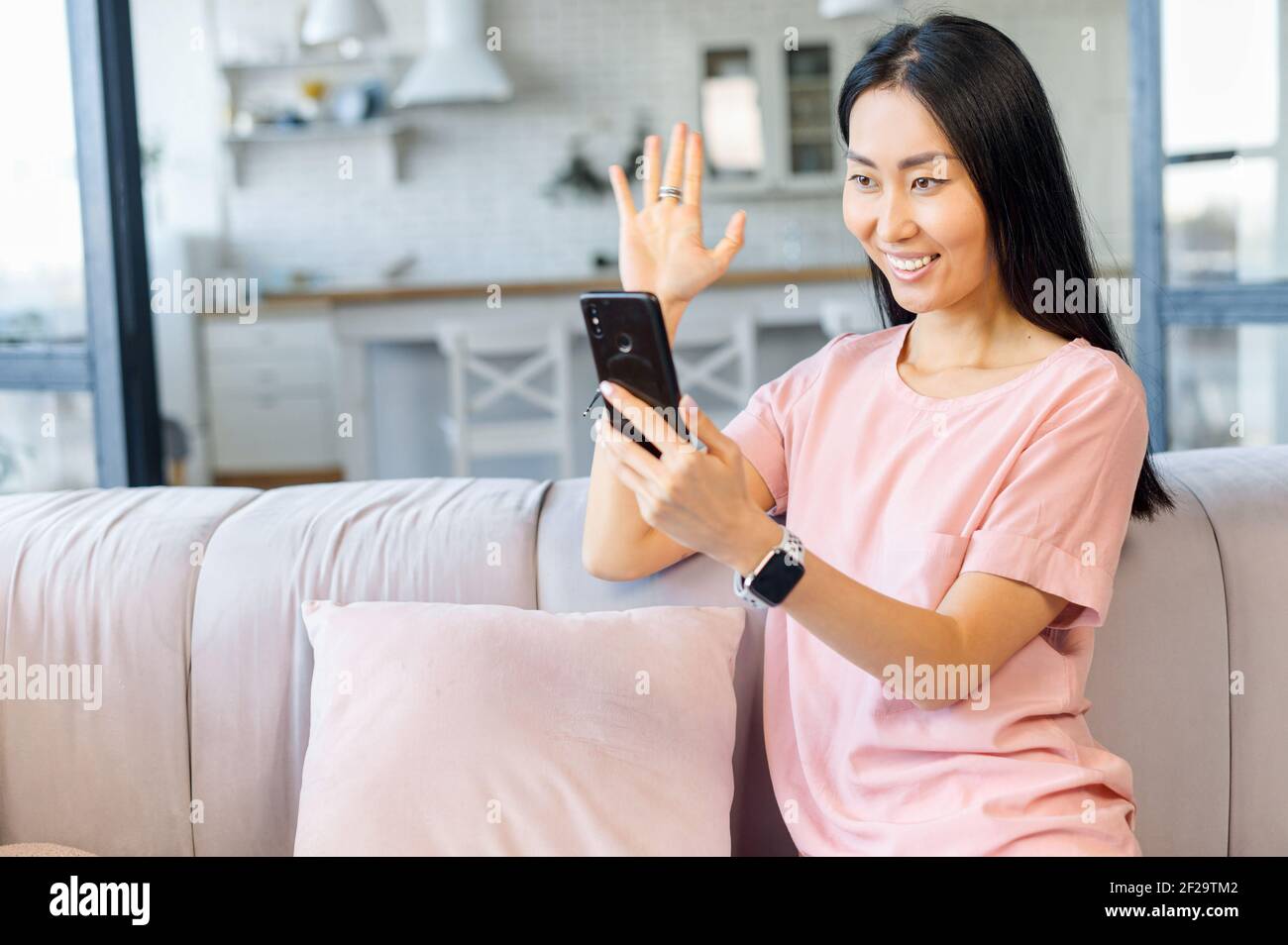 Young Asian mixed-race adult female blogger streaming video online via mobile phone from living room, recording tutorial for social media network, waving, greeting friend with high-five, saying hi Stock Photo photo picture