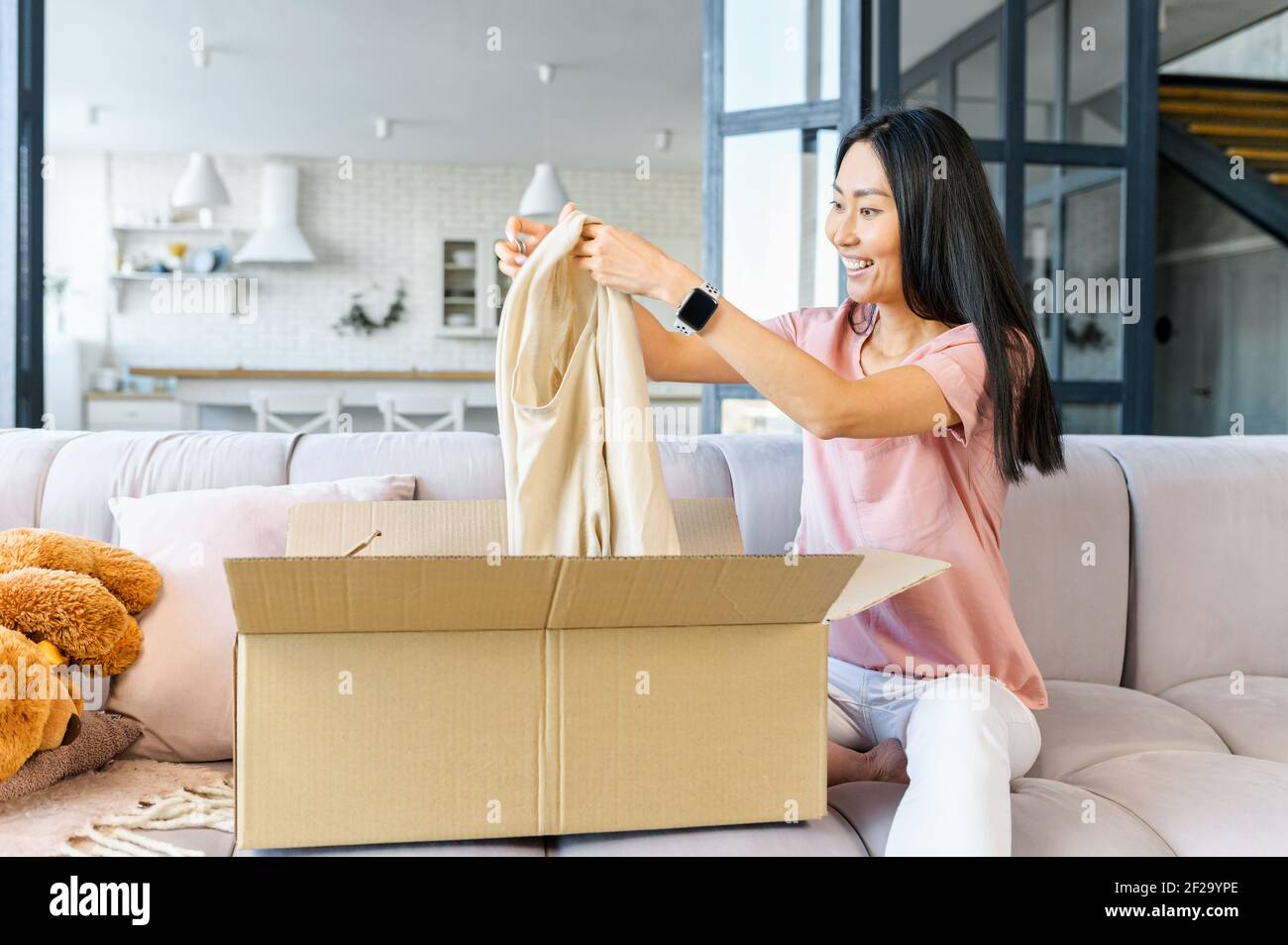 https://c8.alamy.com/comp/2F29YPE/excited-smiling-young-asian-woman-sitting-on-the-couch-holding-received-sweater-from-open-carton-box-unpacking-received-gifts-presents-from-online-store-happy-satisfied-with-fast-delivery-shipment-2F29YPE.jpg