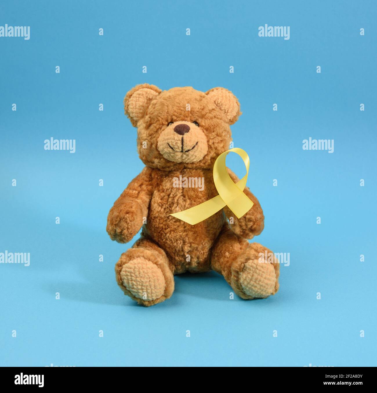 brown teddy bear holds in his paw a yellow ribbon folded in a loop on a blue background. concept of the fight against childhood cancer. problem of sui Stock Photo