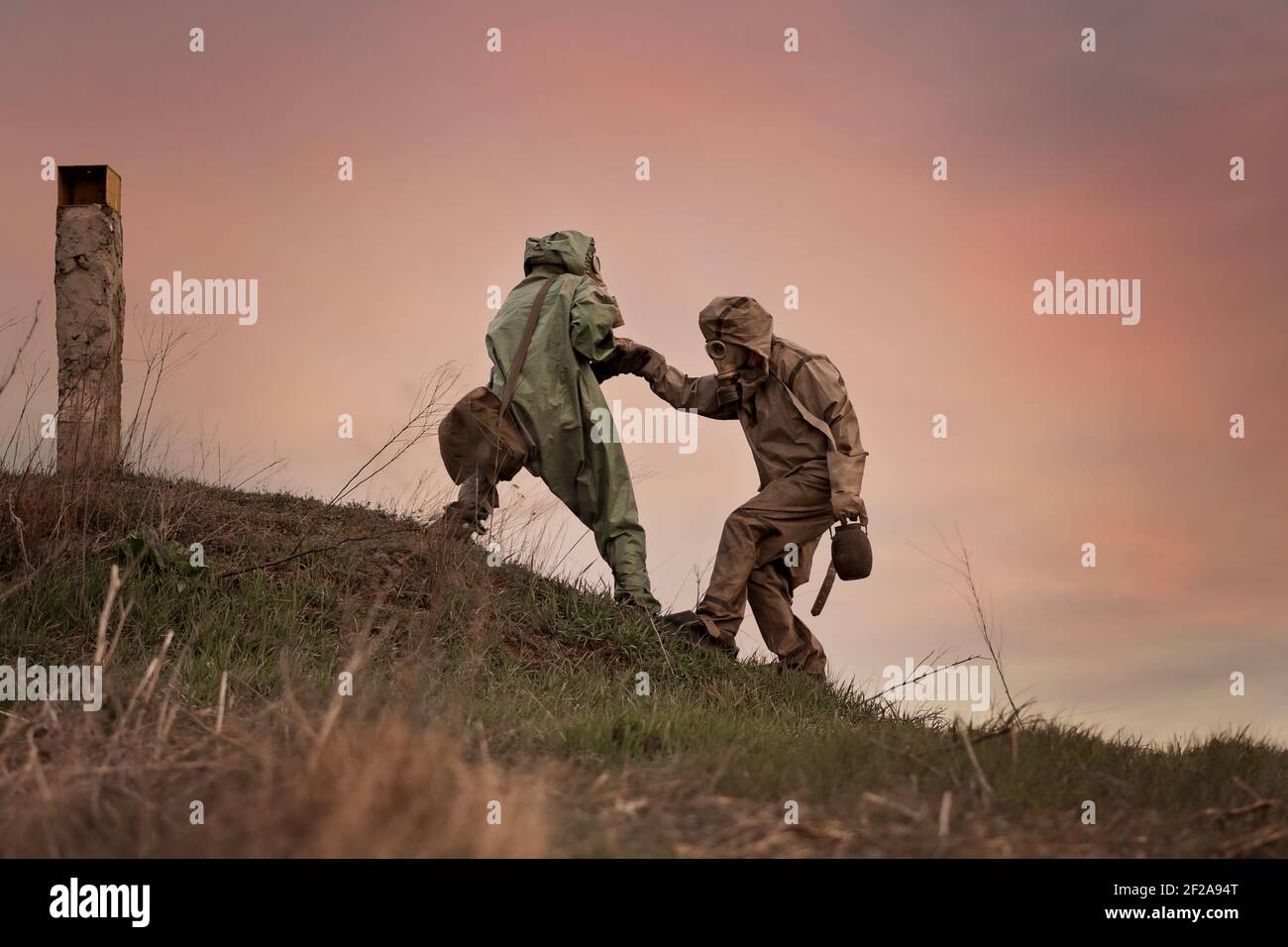 A man in a gas mask and a chemical protection suit helps another person to walk. Post-apocalypse after nuclear war and radiation pollution.  Stock Photo