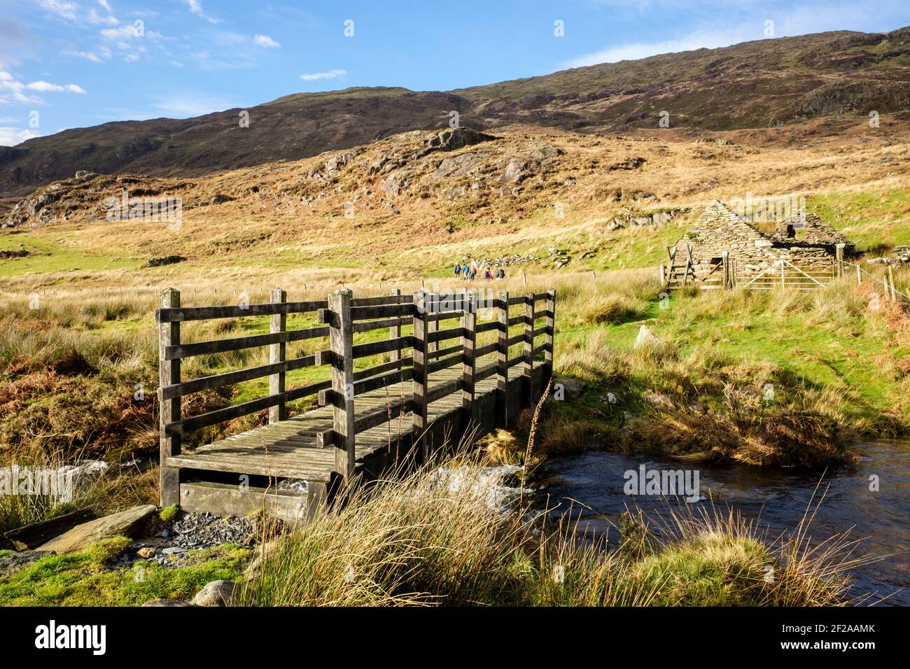 Footbridge crossing over a stream with distant walkers on footpath in Snowdonia National Park. Beddgelert, Gwynedd, Wales, UK, Britain Stock Photo