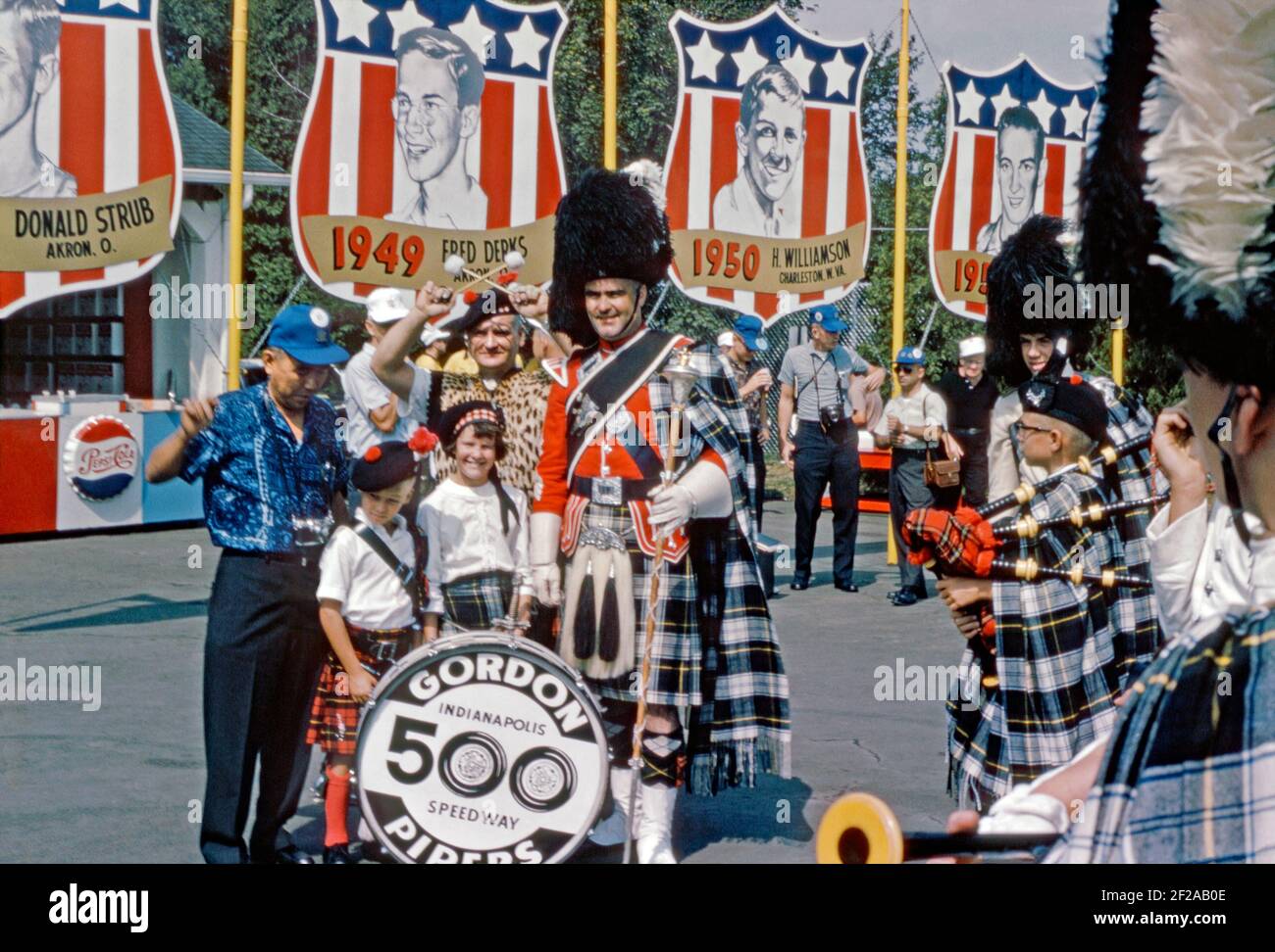 A Scottish pipe band at racetrack at Derby Downs, Akron, Ohio, USA in 1965. The venue has been the home track of the ‘All American Soap Box Derby’ since it was built as a Works Progress Administration (WPA) project in the 1930s. The band members are from the Gordon Pipers from the Indianapolis 500 Speedway. Traditions abound with famous previous winners of races shown on banners, including Fred Derks (1949) and Harold ‘Butch’ Williamson (1950). This image is from an old American amateur Kodak colour transparency – a vintage 1960s photograph. Stock Photo