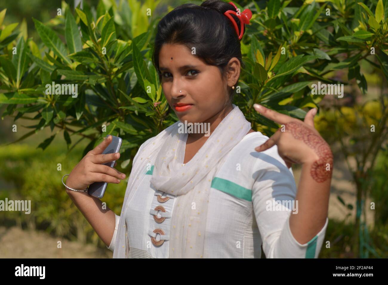 Close up of Indian Bengali teenage girl wearing white salwar, dhupatta, mobile phone on left hand showing victory sign in a park with green leaves Stock Photo