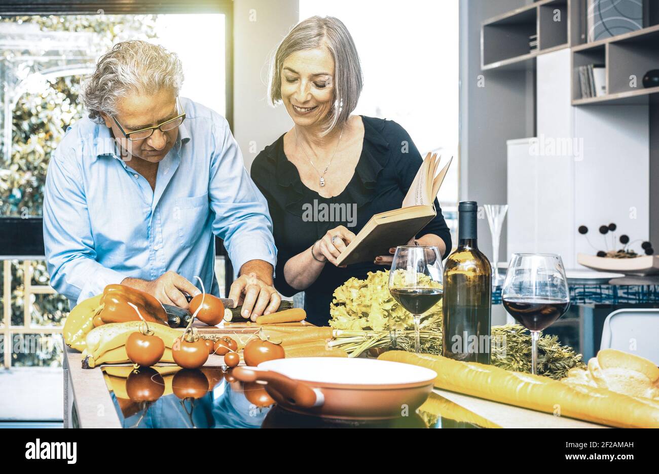 Senior couple cooking healthy food and drinking red wine at house kitchen - Retired people at home preparing lunch with recipe book Stock Photo