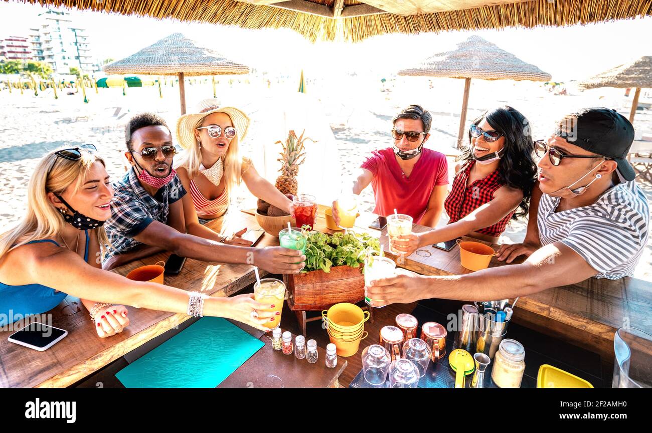 Young trendy people toasting at beach cocktail bar chiringuito with open mask - New normal summer life style concept with friends having fun together Stock Photo