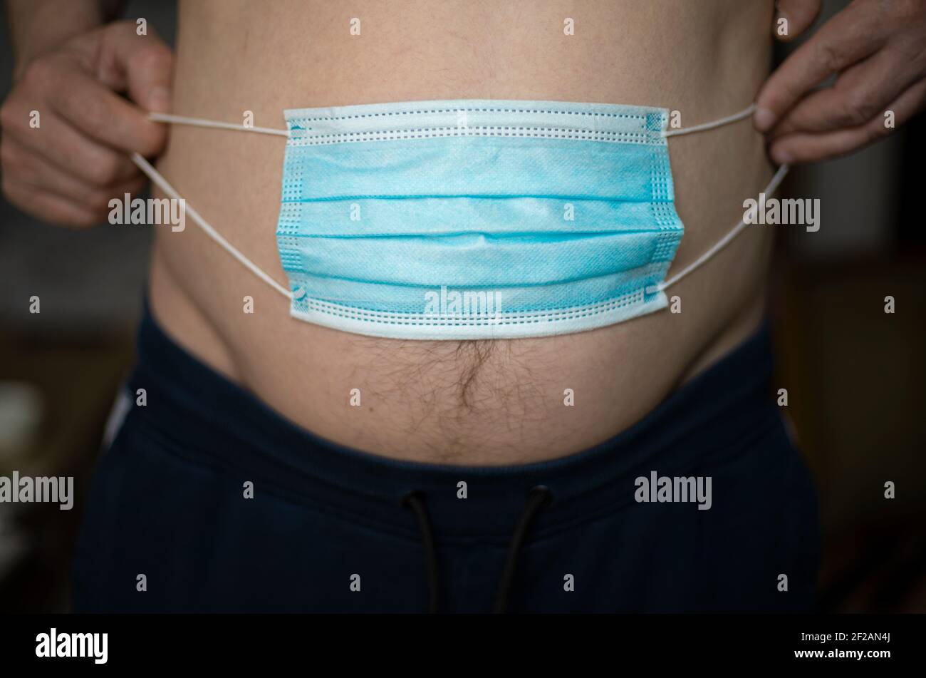 A man holding a blue medical mask over his stomach will gain weight during the pandemic of the coronavirus crisis. Obesity concept Stock Photo