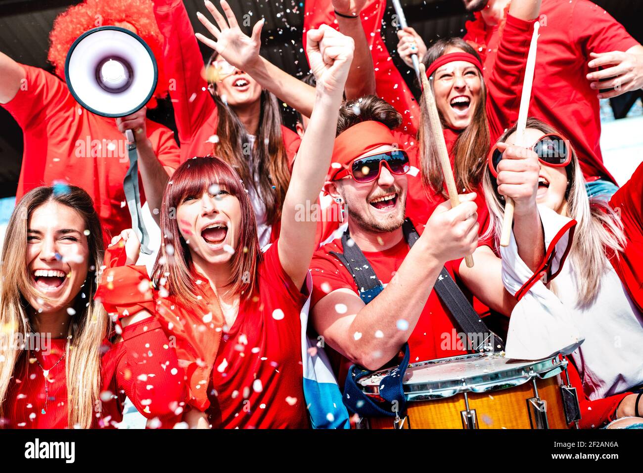 Football supporter fans cheering with confetti watching soccer match cup at stadium tribune - Young people group with red t-shirt having excited fun Stock Photo