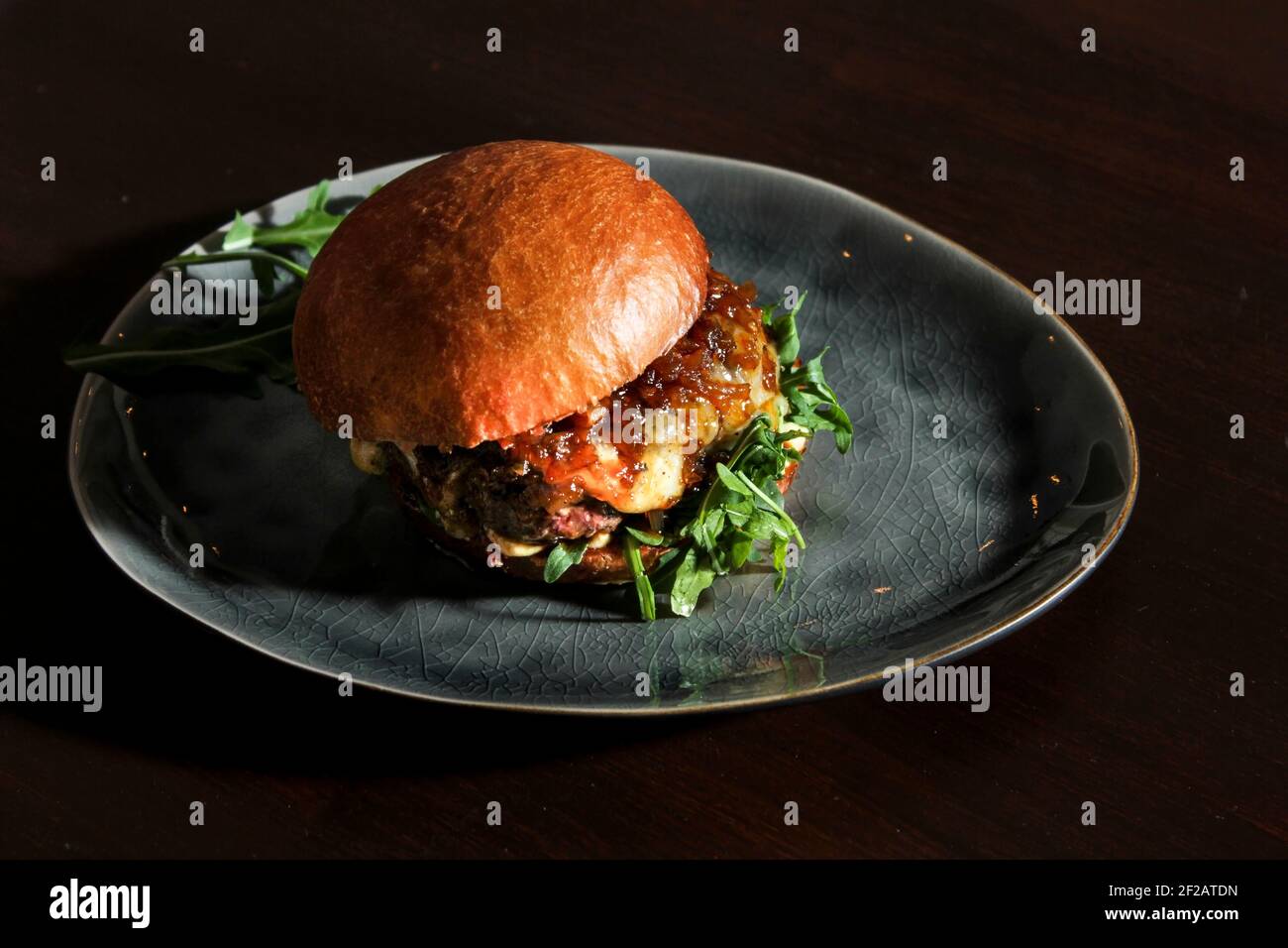 Juicy beef burger with caramelized onion, cheese and rucola on homemade bun. Hamburger served on vintage handmade grey plate on the dark brown table. Stock Photo