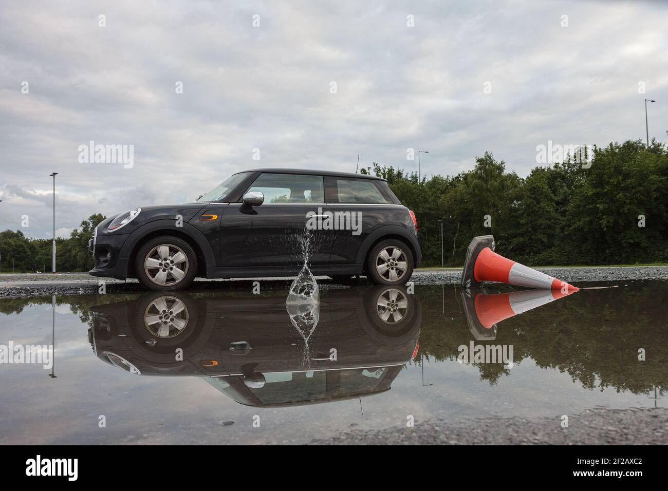 Mini Cooper & Overturned Road Cone in the Puddle, car reflection in water, cloudy sky, green trees in the background, plus overturned traffic cone Stock Photo