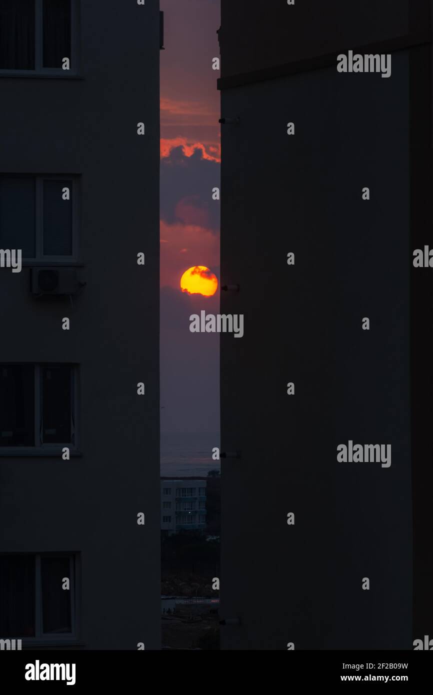 The setting orange round sun sets between two houses. Twilight in the city. Fog over the sea and a beautiful sunset. The concept of urban life, evenin Stock Photo