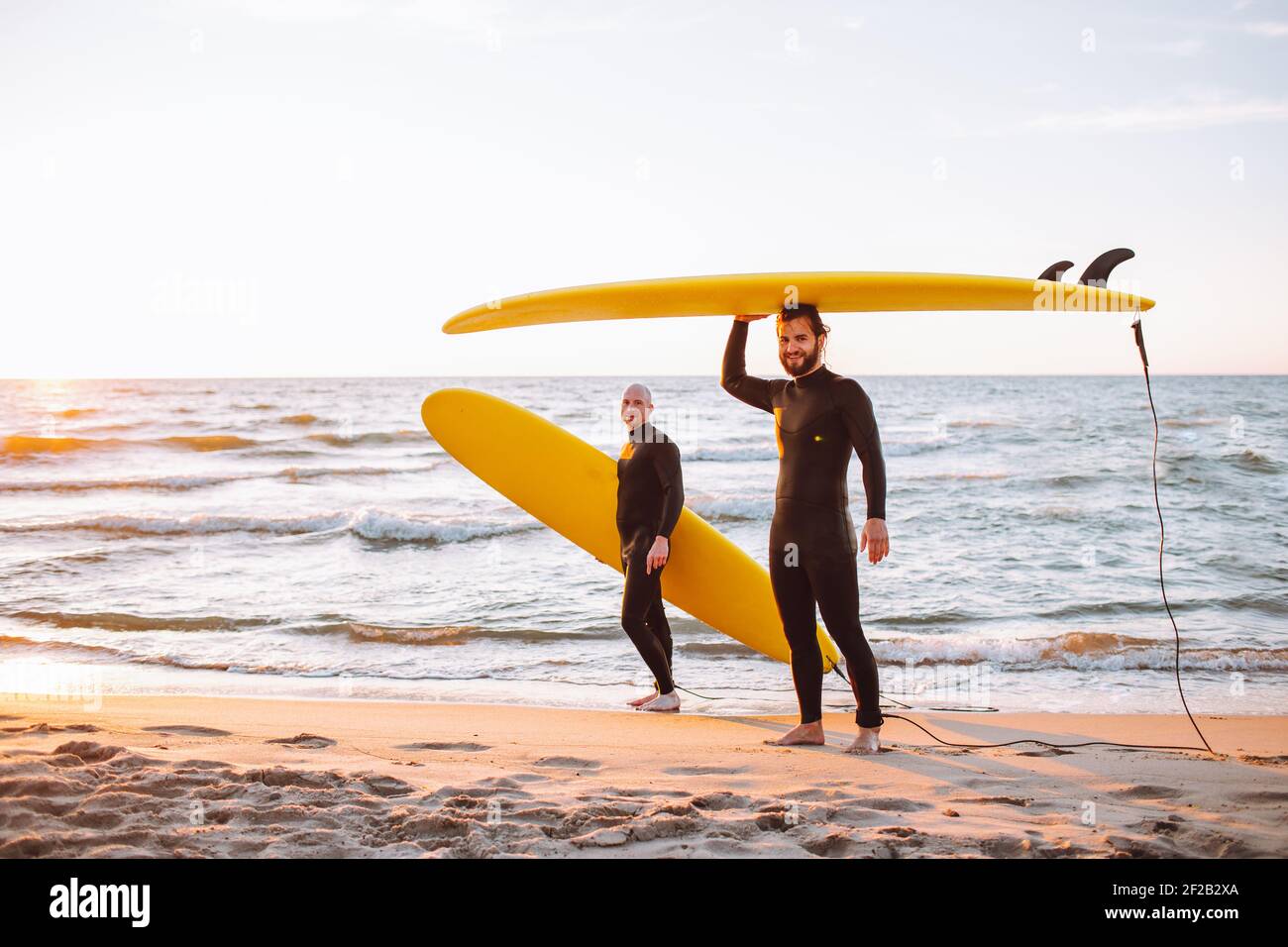 Two young surfers in black wetsuit with yellow surfing longboards at ocean coast at sunset
