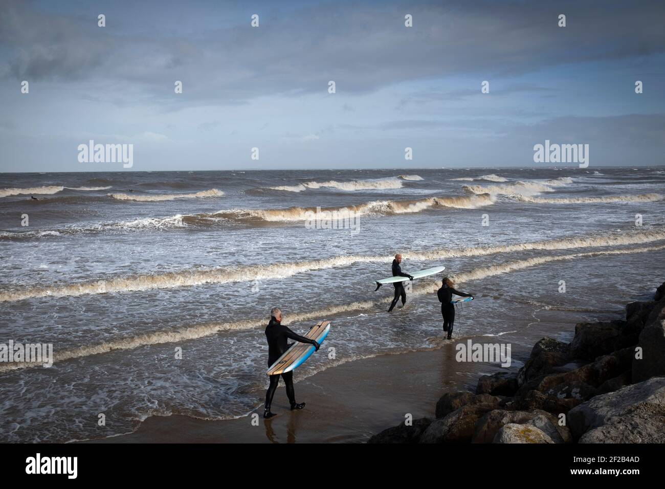 Three surfers walking towards the sea in the mouth of the river Mersey. High tides and a storm which swept across the United Kingdom brought favourable conditions for surfing in Leasowe Bay, Merseyside on 11 March, 2021. Stock Photo
