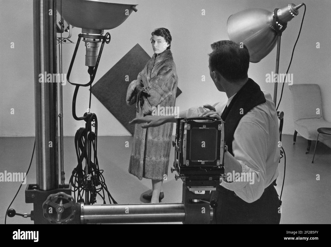 1950s fashion photographer and model. A young woman is dressed up the 1950s fashionable fur coat and is being instructed by the photographer by the camera. Lights and background is arranged. Sweden august 1957 Stock Photo