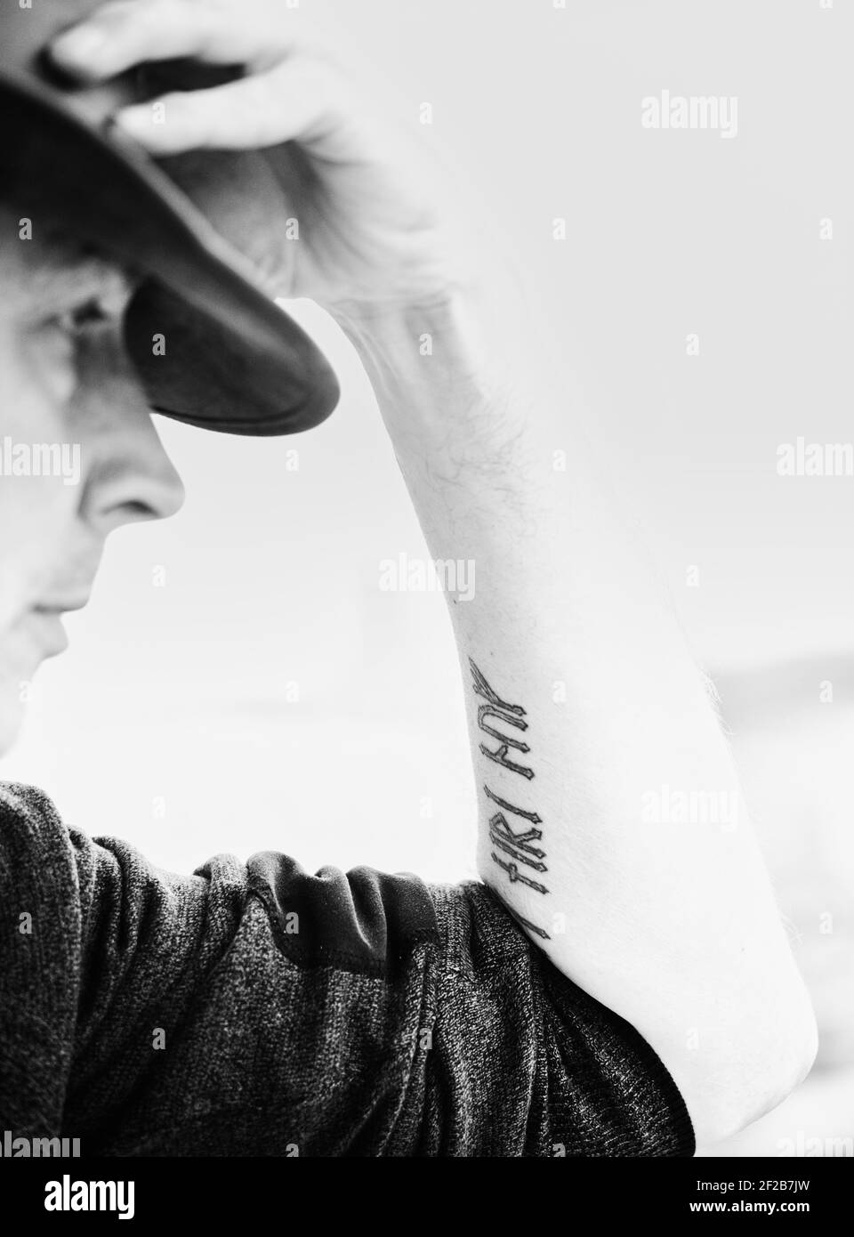Black and white side view image of serious mid adult man with hand on hat and arm tattoo. Concept of lifestyle, individuality Stock Photo