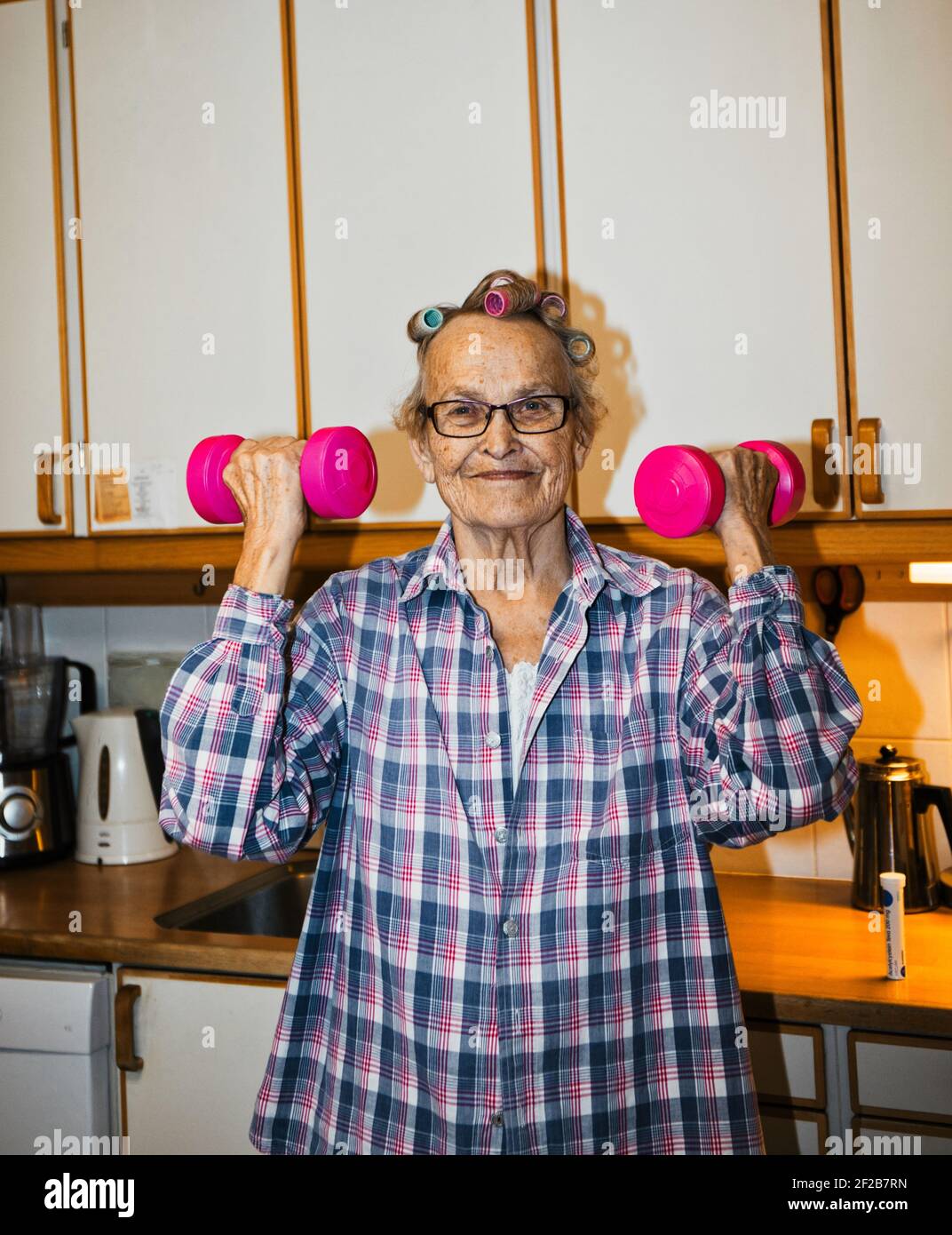 Cheerful senior elderly Swedish woman lifting pink dumbbells in kitchen. Concept of lifestyle, seniors exercising, active lifestyle Stock Photo