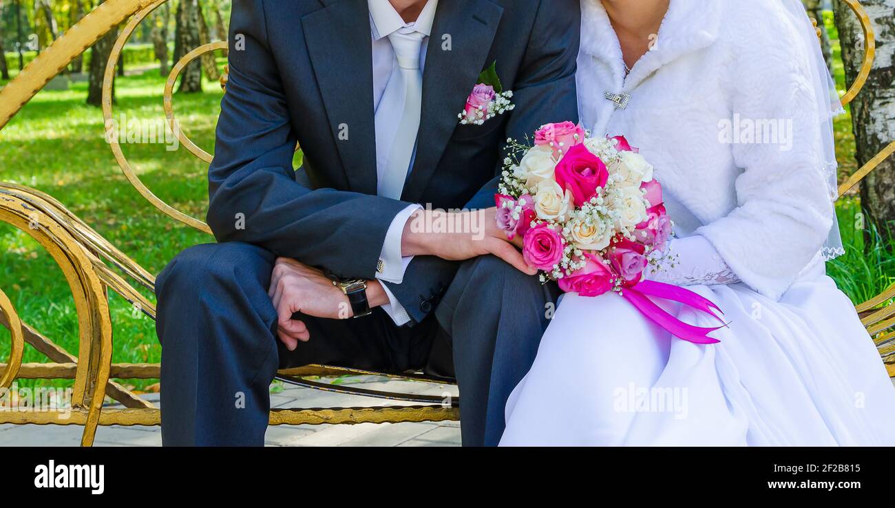 The groom and the snow-white bride are sitting on a yellow bench and holding a wedding bouquet of multi-colored roses in front of them. Stock Photo