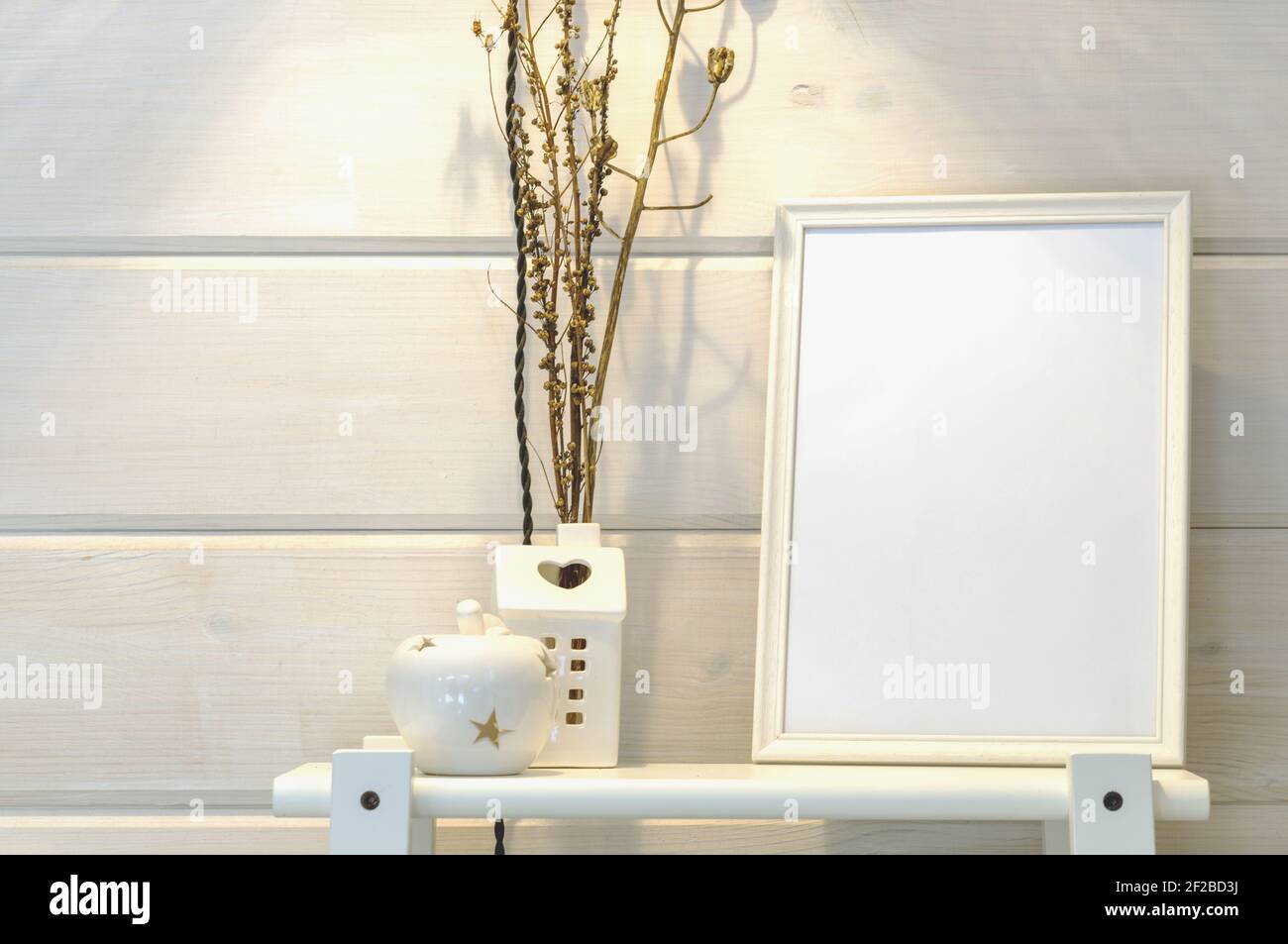 Wooden white wall in a scandinavian house, mockup of a white photo frame on a shelf, dry flowers. Stock Photo