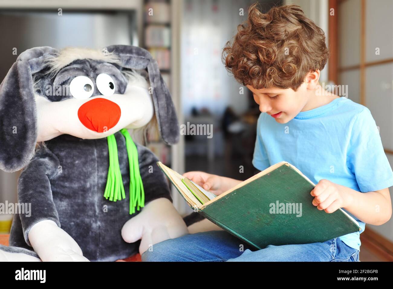 Adorable little boy with curly hair reads an exciting, fun and interesting book to his plush rabbit and smiles Stock Photo