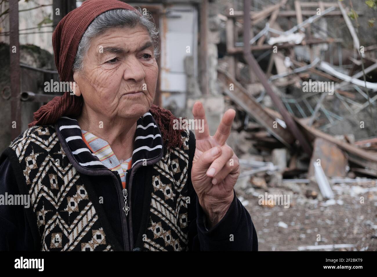 An elderly woman gives the V-sign in front of a house destroyed by Azerbaijan's artillery during a military conflict in Stepanakert the de facto capital of the self-proclaimed Republic of Artsakh or Nagorno-Karabakh, de jure part of the Republic of Azerbaijan on November 06, 2020. The fighting between Armenia and Azerbaijan over Nagorno-Karabakh known also as the Artsakh Republic re-erupted in late September into a six-week war with both countries accusing each other of provocation that left thousands dead. Stock Photo