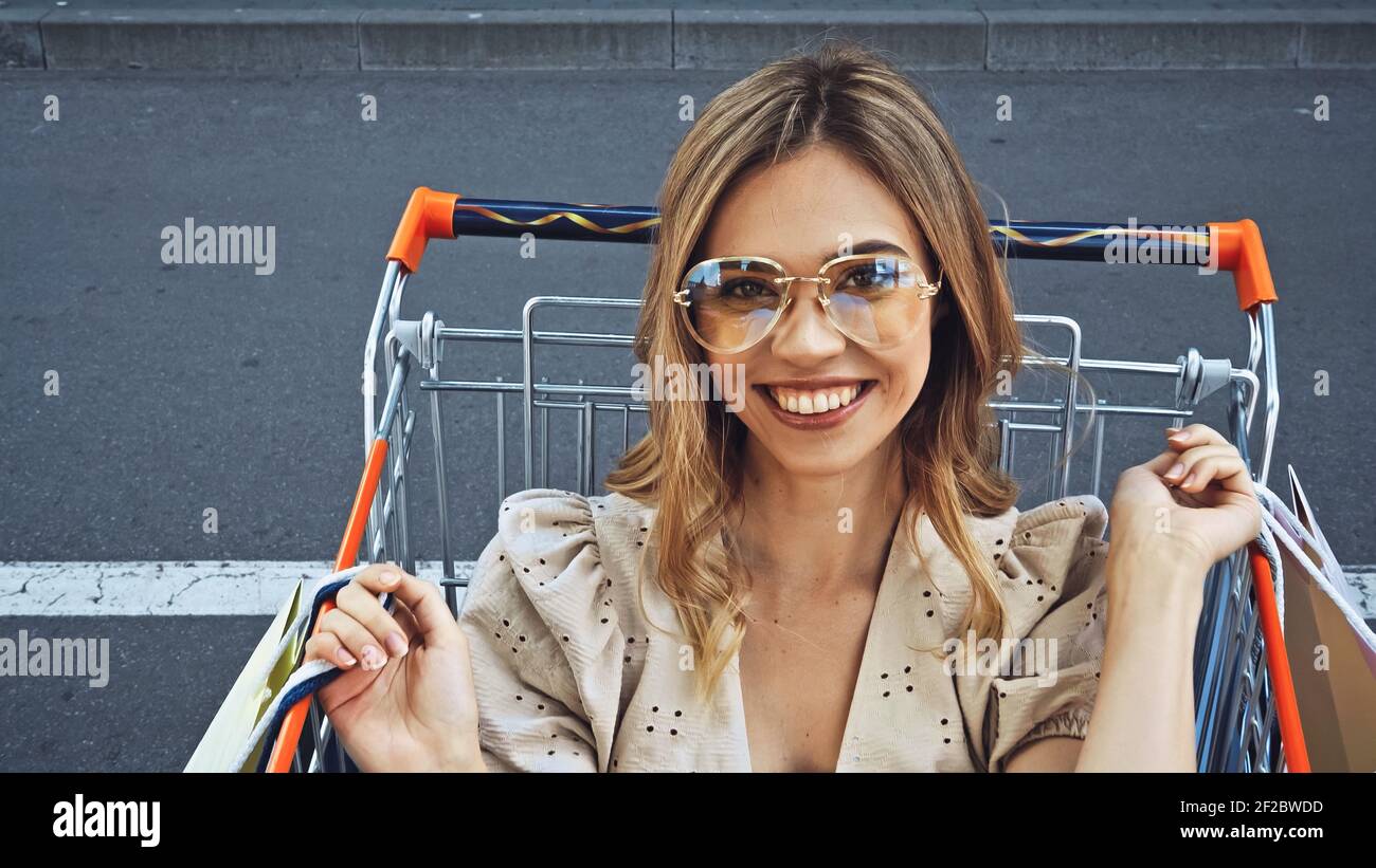 happy woman holding shopping bags while sitting in cart Stock Photo - Alamy