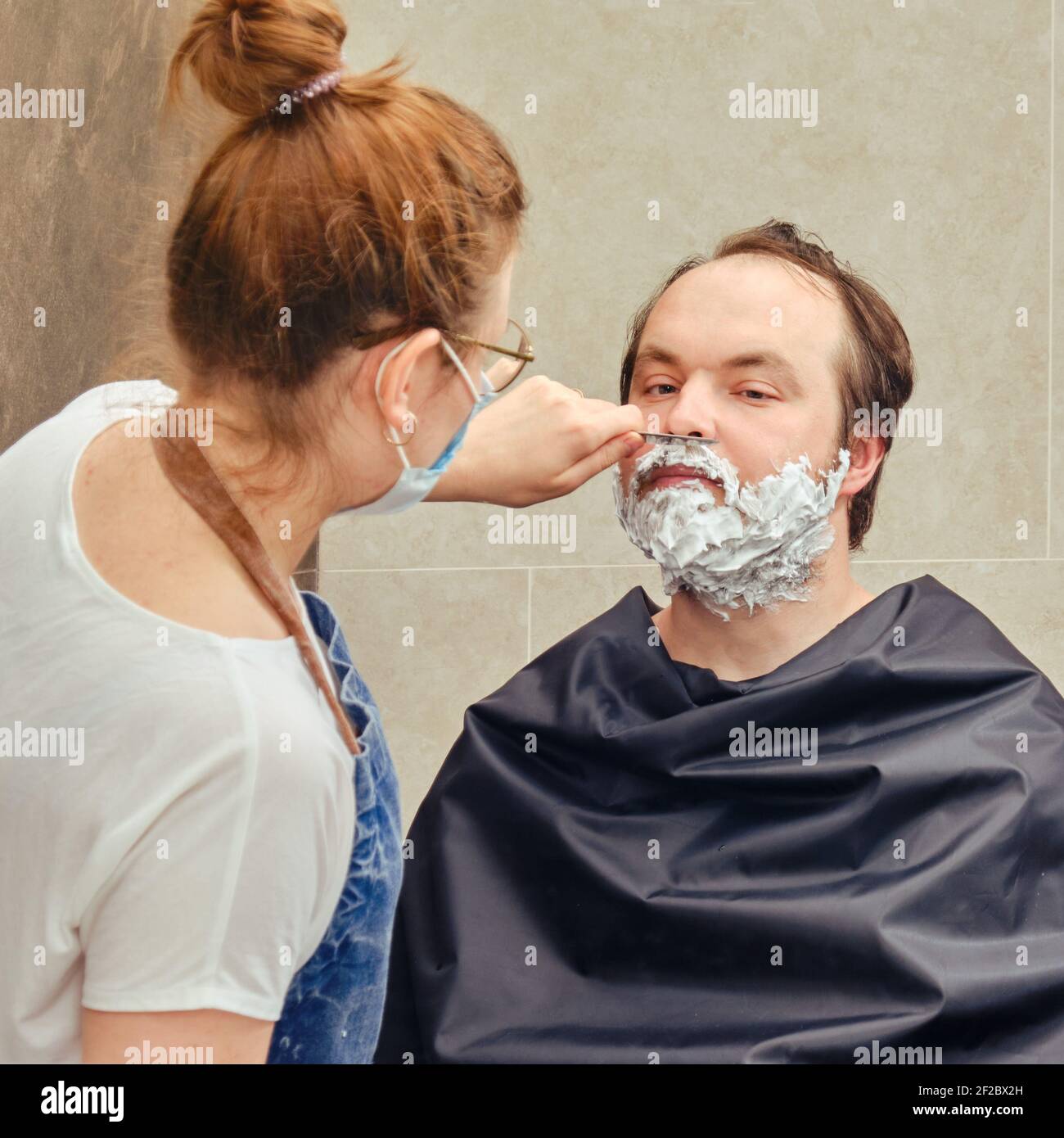 A woman shaves a man mustache with a straight razor. Concept of closed hairdressers and barbershops in isolation from the epidemic of the coronavirus Stock Photo