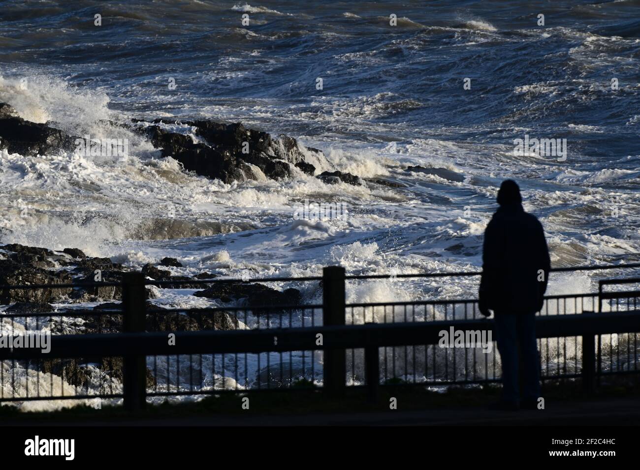 Strong Winds and Storm Surf batter the coastline at Mumbles Head. A walker is stopped in his tracks in awe of the power of the waves. Stock Photo