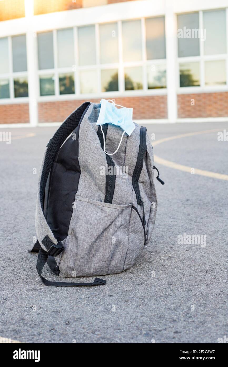 School backpack with face mask on the road near school building. Back to school during epidemic concept. Stock Photo