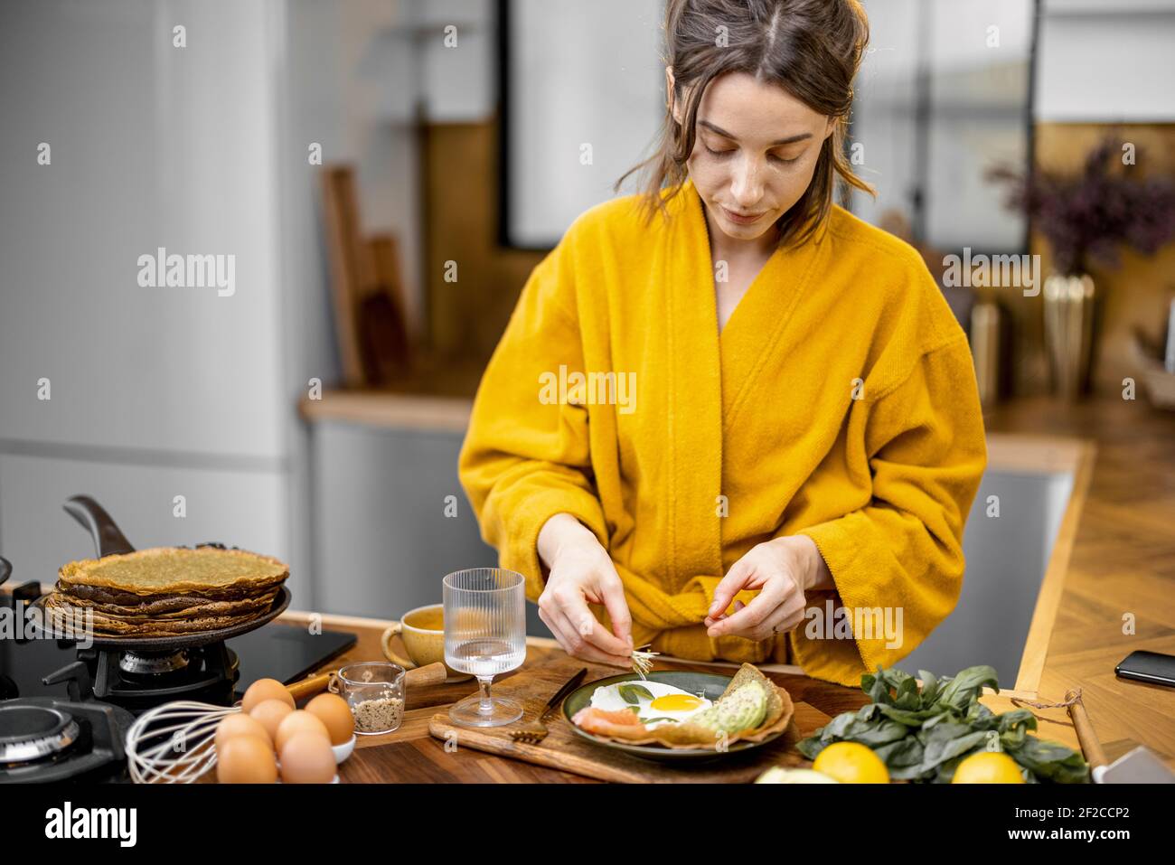 Young woman dressed in yellow bathrobe preparing eggs for breakfast, putting greens on a plate on the kitchen at home Stock Photo