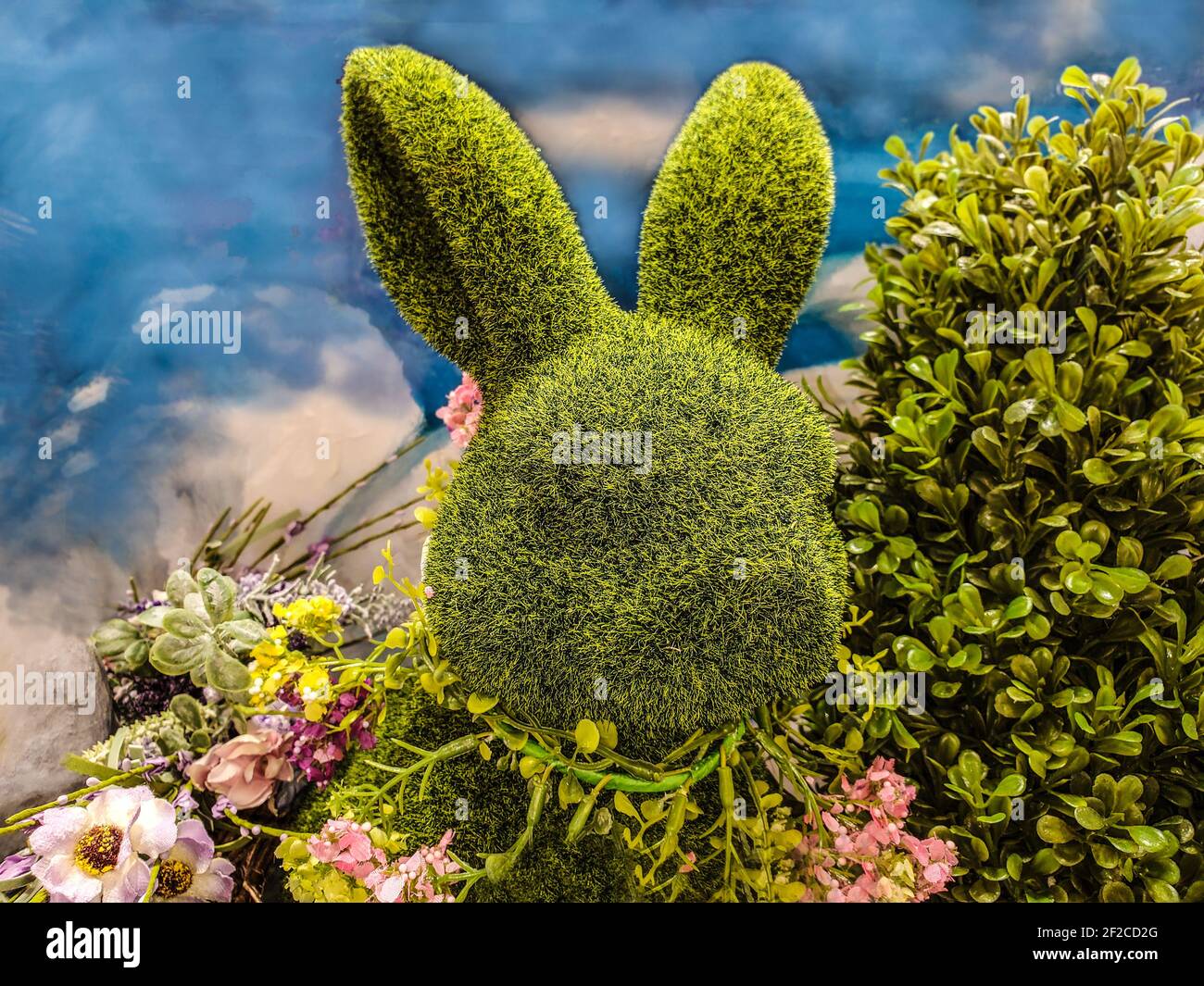 Decorative green topiary rabbit head with flowers around its neck and surrounded by greenery in front of blue sky-like background - Great for spring o Stock Photo