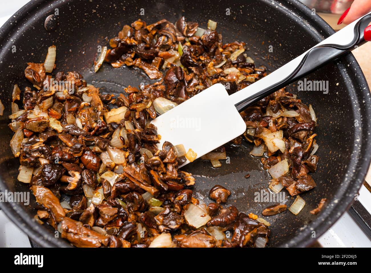 A woman is holding a silicone spoon and mixing fried mushrooms with onions in a frying pan on a