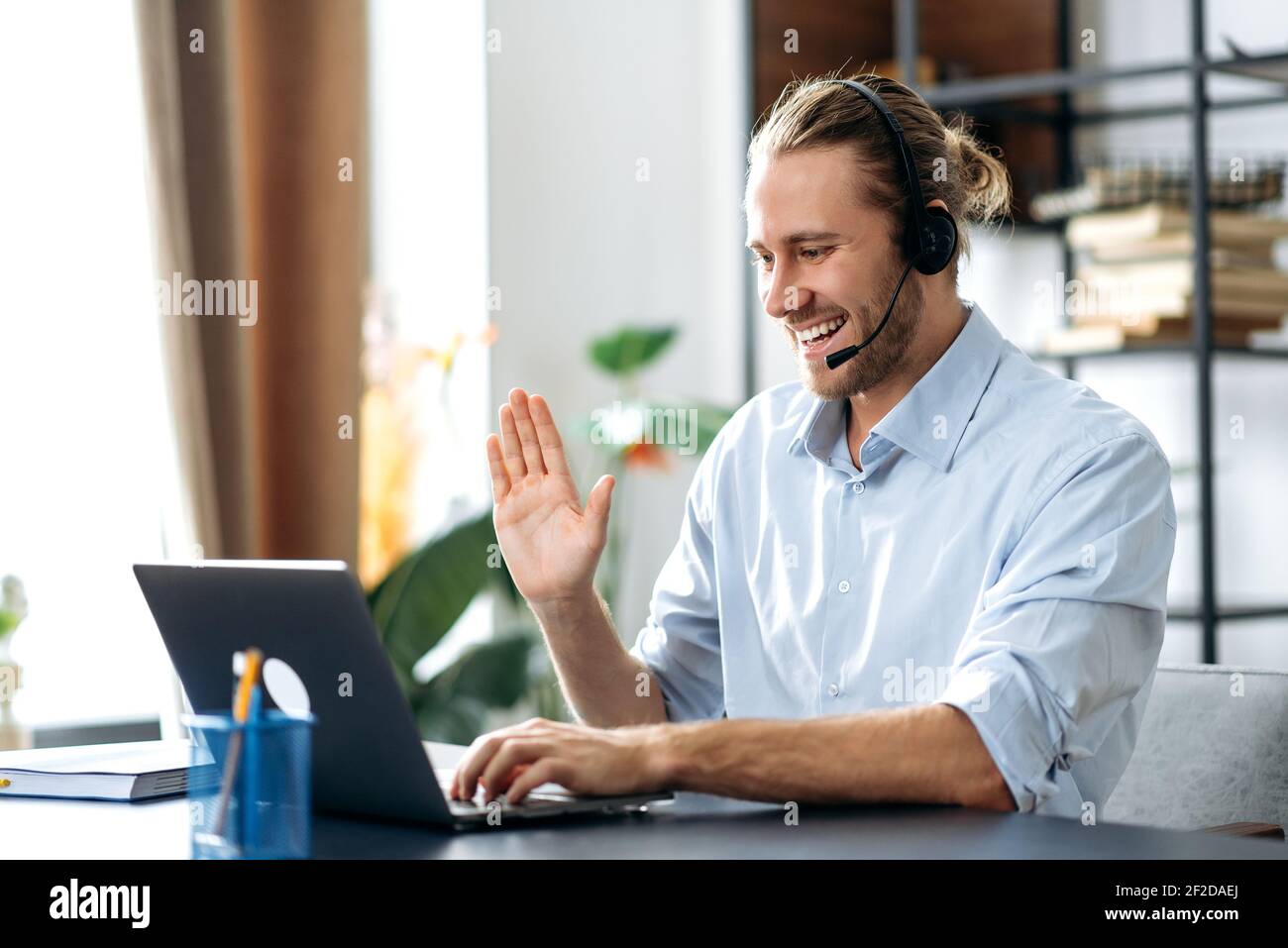Joyful freelancer guy with headset, uses laptop, have a video call, communicate with colleagues or friends by video conference, waving hand and smiling. Handsome caucasian man working from home Stock Photo