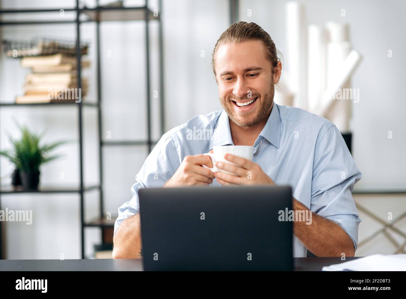 Happy confident caucasian guy, employee or freelancer, took a break from work, holds a cup of coffee while sitting at his desk at home or office, looking at the laptop with a friendly smile Stock Photo
