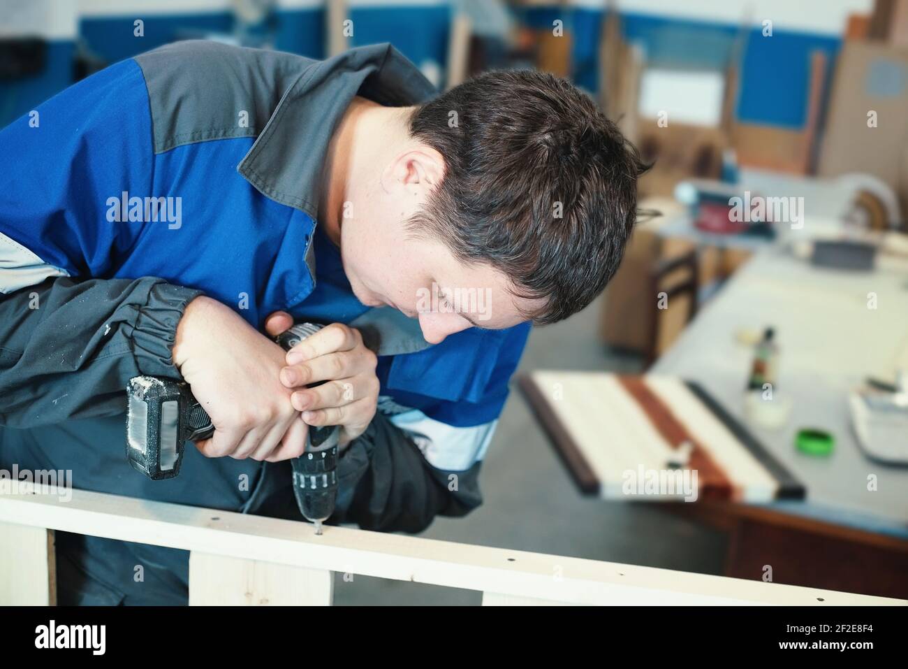 A young worker in overalls with a screwdriver collects furniture in a carpenter's shop. Background for making furniture. Stock Photo