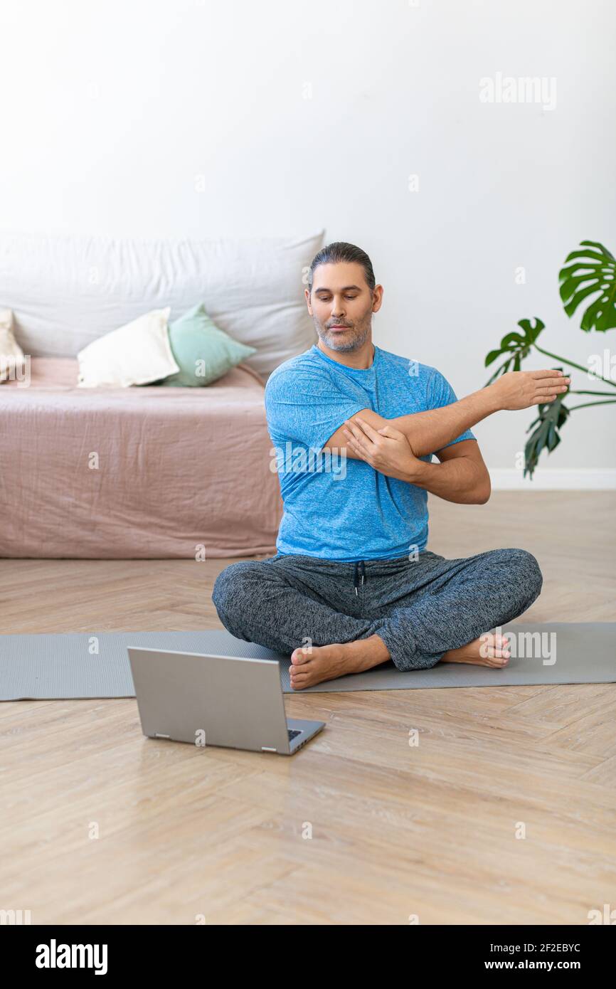 Vertical photo of a middle aged man during online steaming. Home workout - stretching muscles before workout. A man practicing pilates online. Vertica Stock Photo