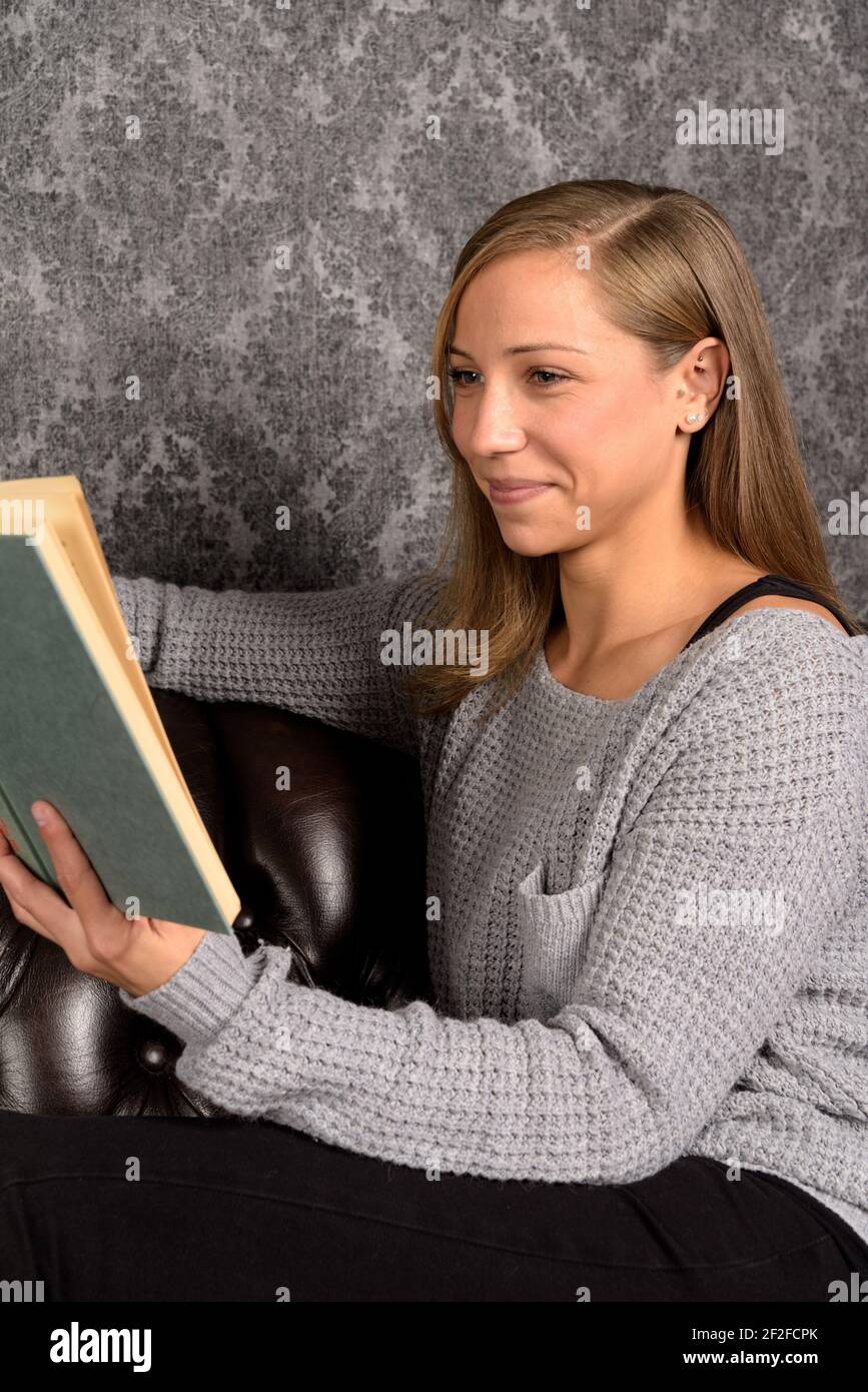 Relaxed european woman in woolen pullover reading a book and smiling while sitting on a leather sofa Stock Photo