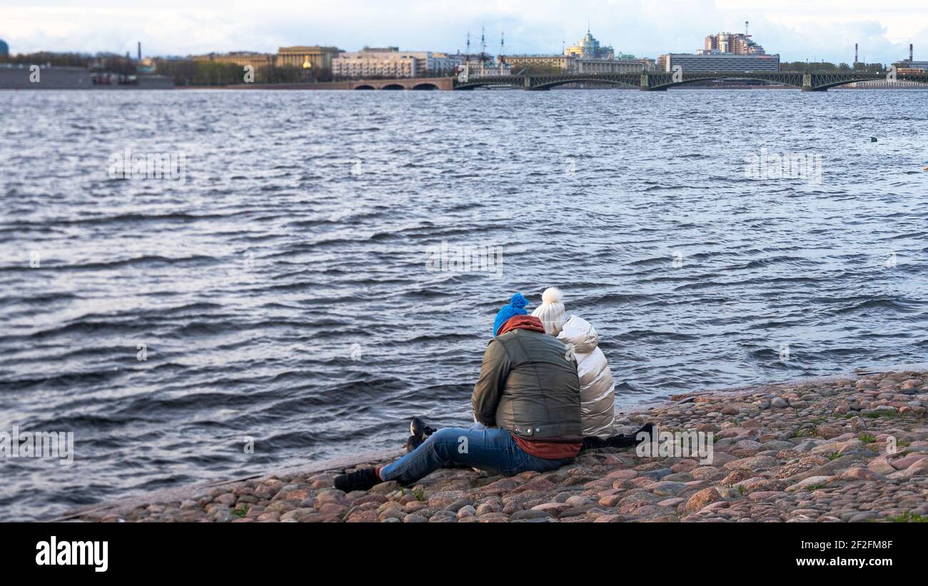 Two persons in warm clothing are sitting on the cobbles of the bank of the river, backview Stock Photo