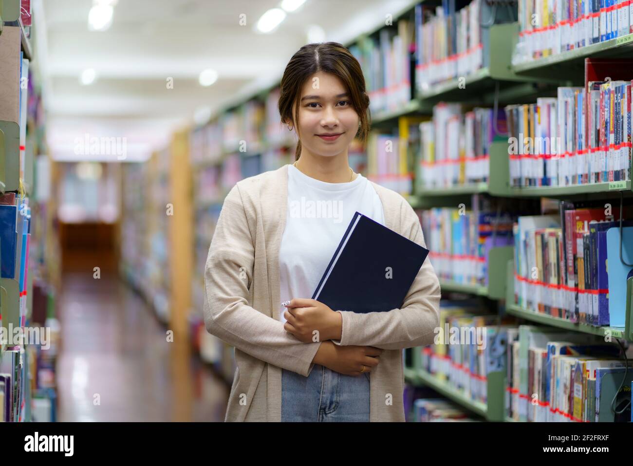 Portrait of Smart Asian woman university student reading book and looking at camera between bookshelves in campus library with copyspace. Stock Photo