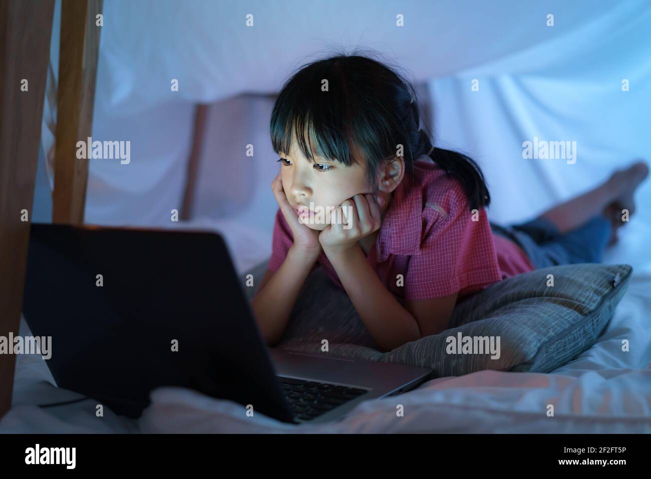 Asian girl child to make a camp to play imaginatively watching a film on laptop in the darkness of the camp in living room at home. Stock Photo