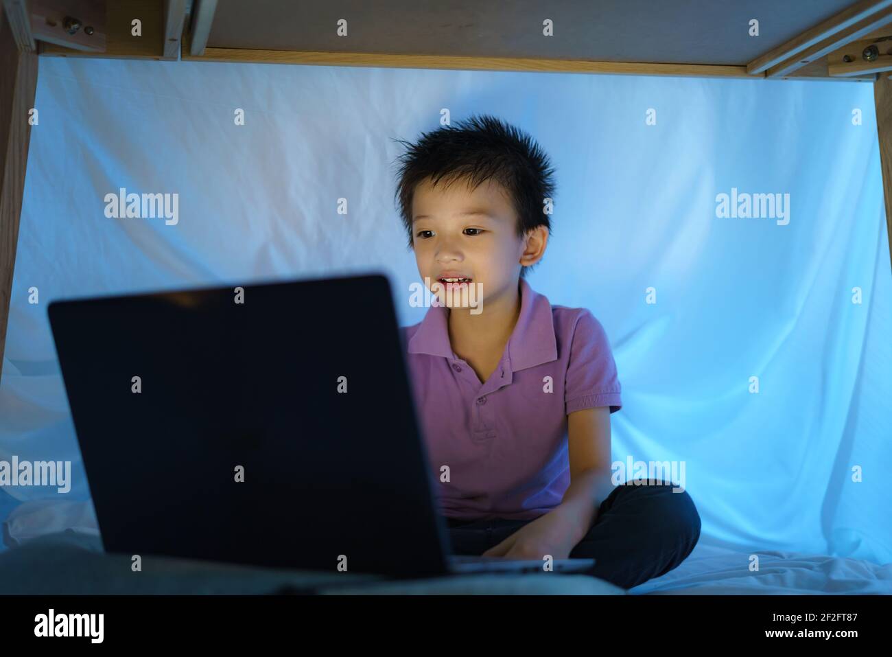 Asian boy child to make a camp to play imaginatively watching a film on laptop in the darkness of the camp in living room at home. Stock Photo